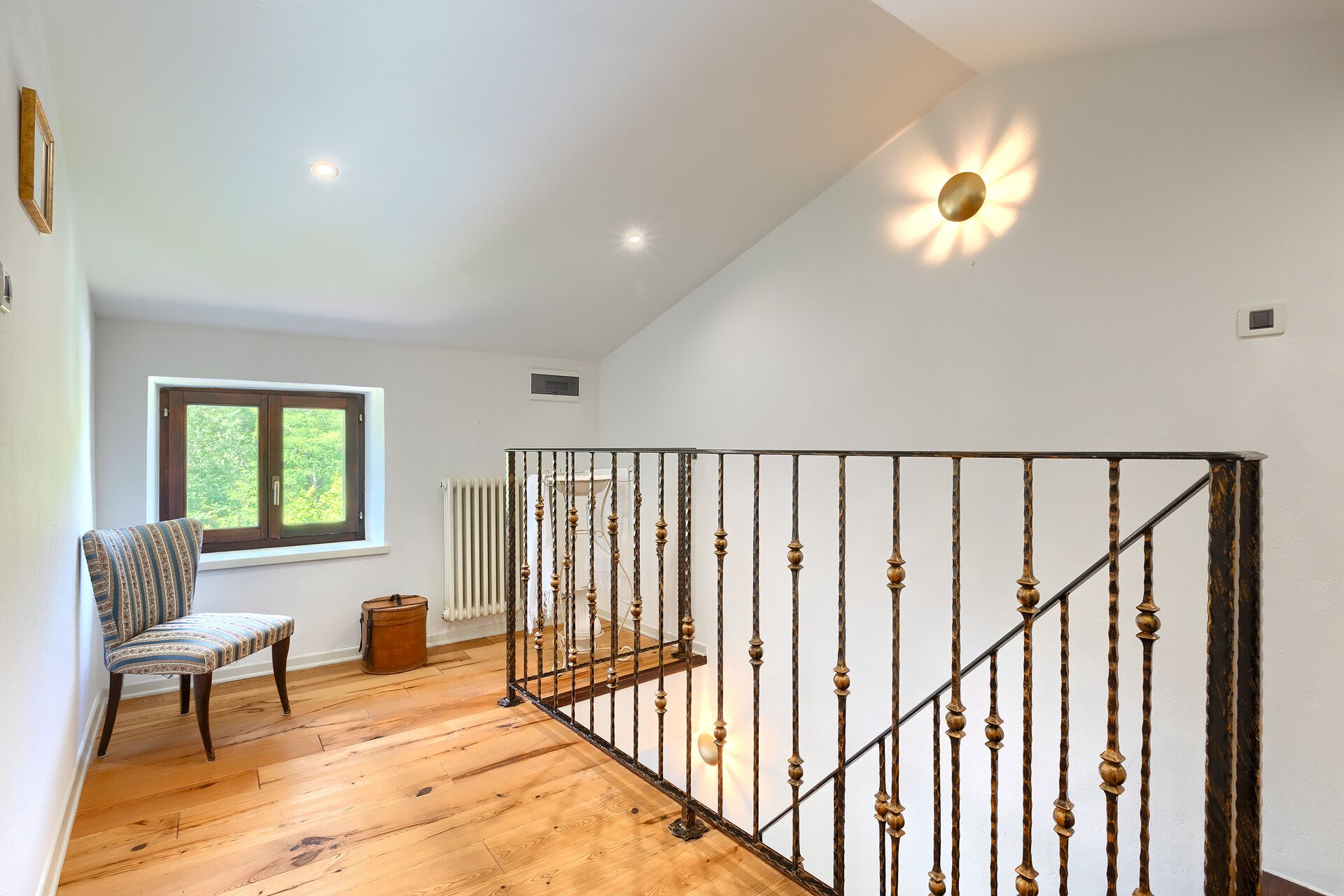A staircase with a metal railing, a wooden floor, a chair, a window with a view of greenery, and a radiator.