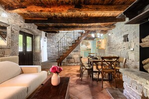 Un intérieur rustique avec des poutres apparentes, des murs en pierre, un coin repas avec des meubles en bois et un escalier menant à un niveau supérieur.
