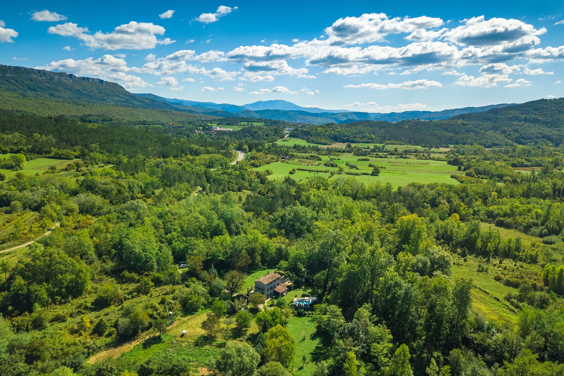A rural landscape with rolling hills, a winding road, and a small house surrounded by greenery.