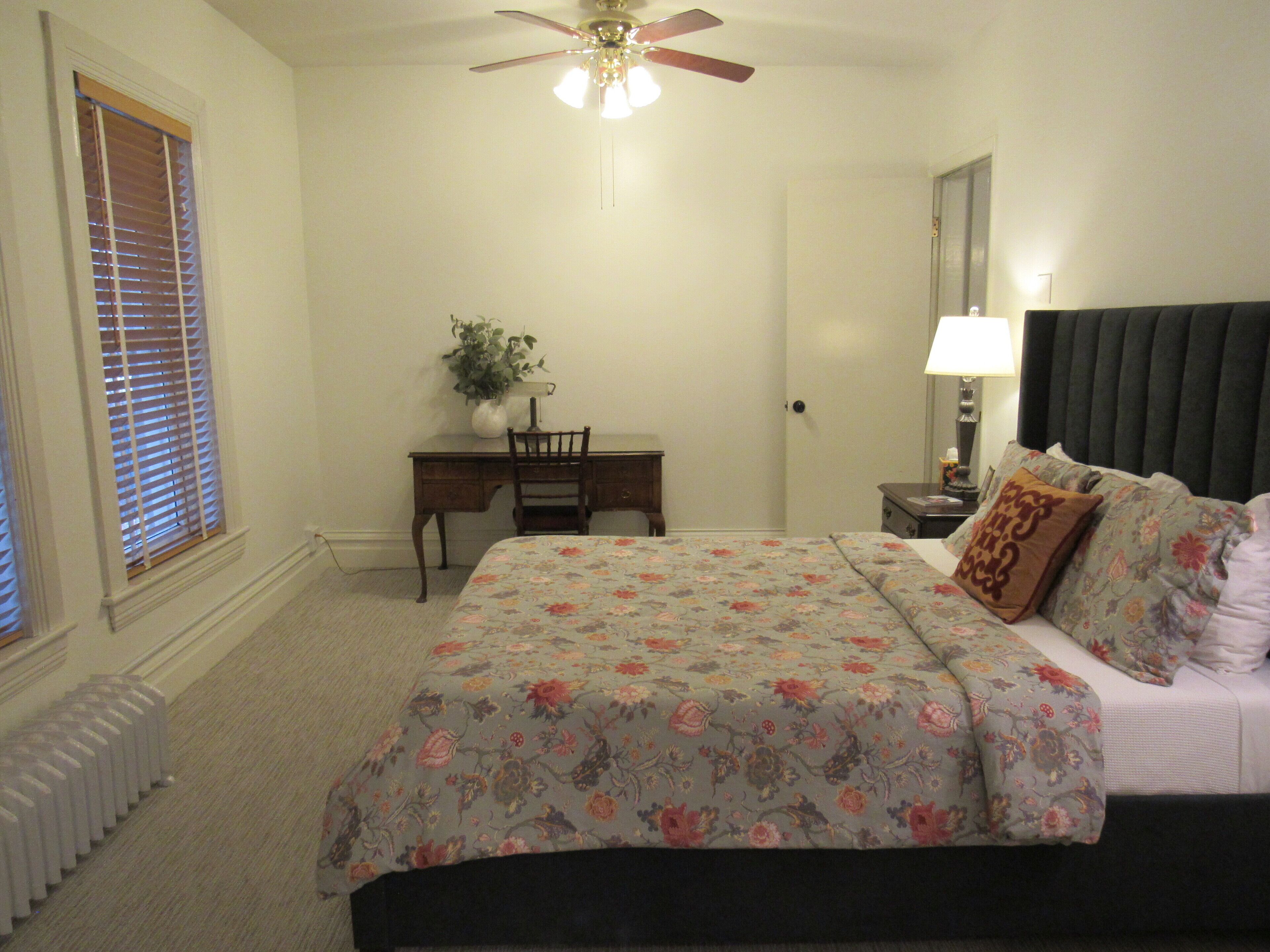 A bedroom with a bed, a desk, a chair, a ceiling fan, and a radiator.