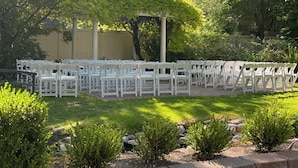 A paved outdoor area with white chairs arranged in rows, surrounded by greenery and a brick wall. - The Riter Mansion (Logan)