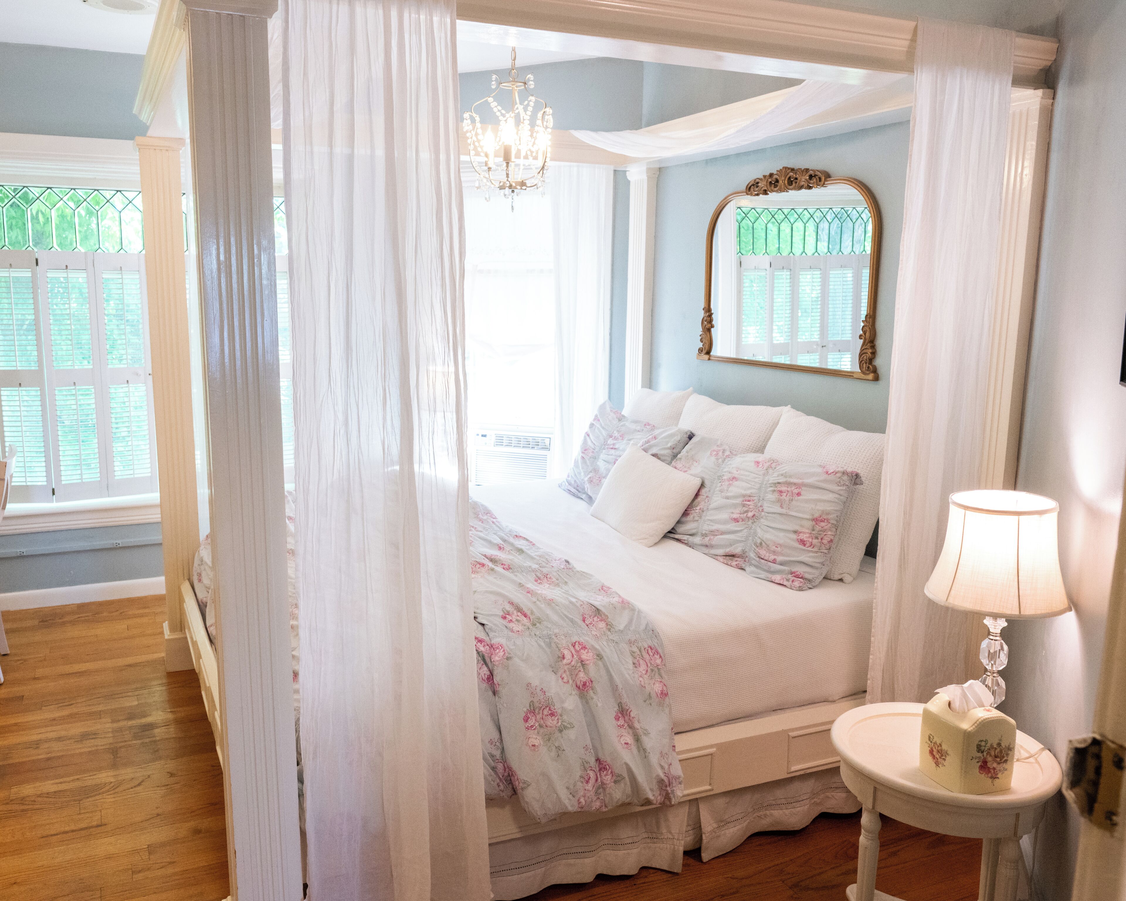 A four-poster canopy bed with floral bedding, a bedside lamp, and a mirror on the wall.