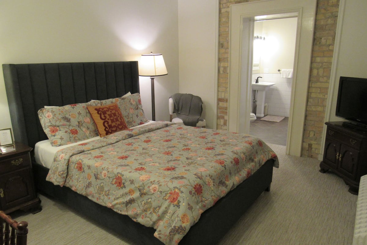 A bedroom with a bed, a nightstand, a lamp, a chair, a television, and a bathroom visible through an open door.