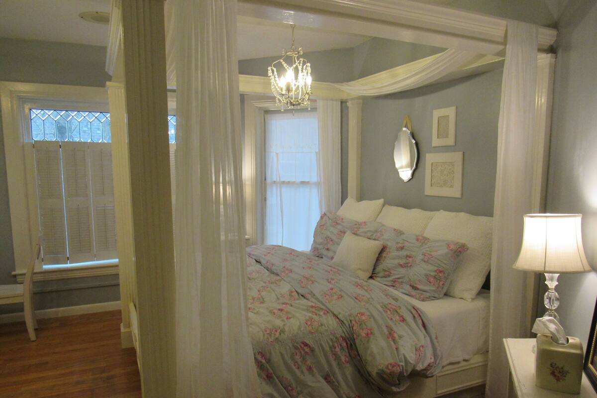 A bedroom with a canopy bed, a chandelier, a bedside table with a lamp, and a window with shutters.