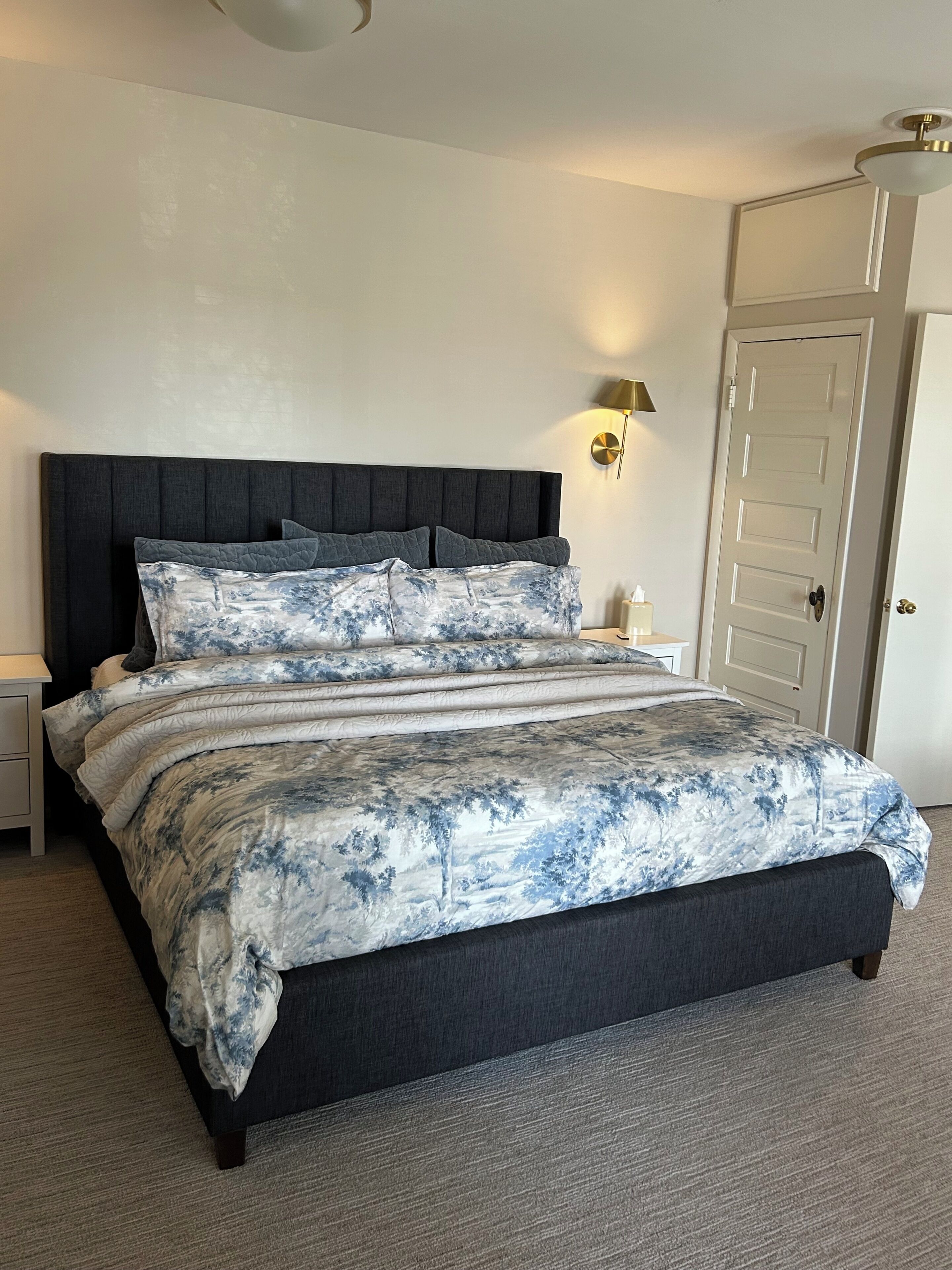 A bedroom with a dark headboard, a bed with a blue and white floral bedspread, a bedside table, a wall-mounted lamp, and a door.