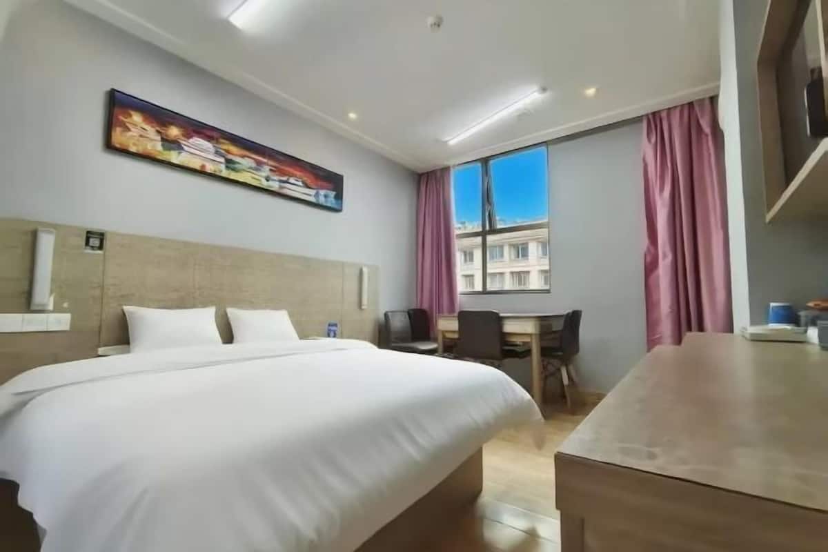 A hotel room with a bed, a desk, a chair, a window with curtains, and a painting on the wall.
