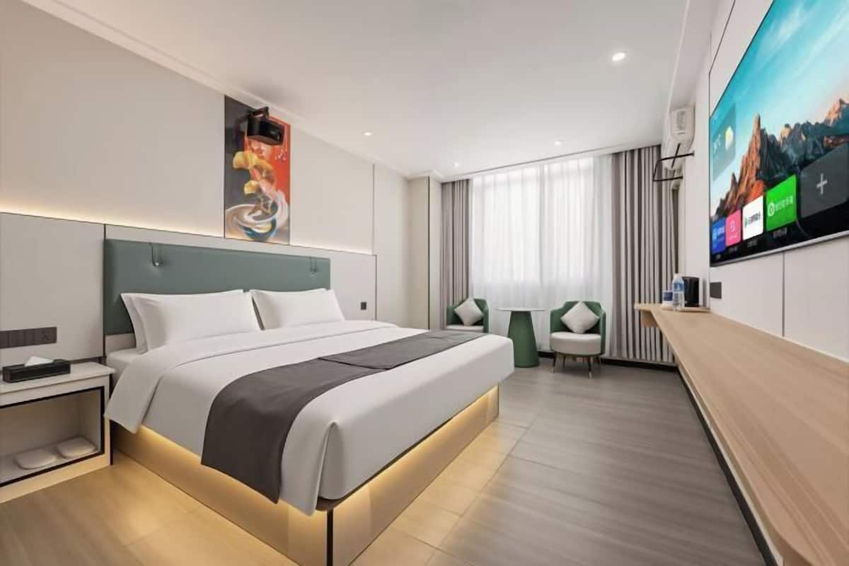 A modern hotel room with a large bed, a TV mounted on the wall, a desk, and a seating area with a chair and table.