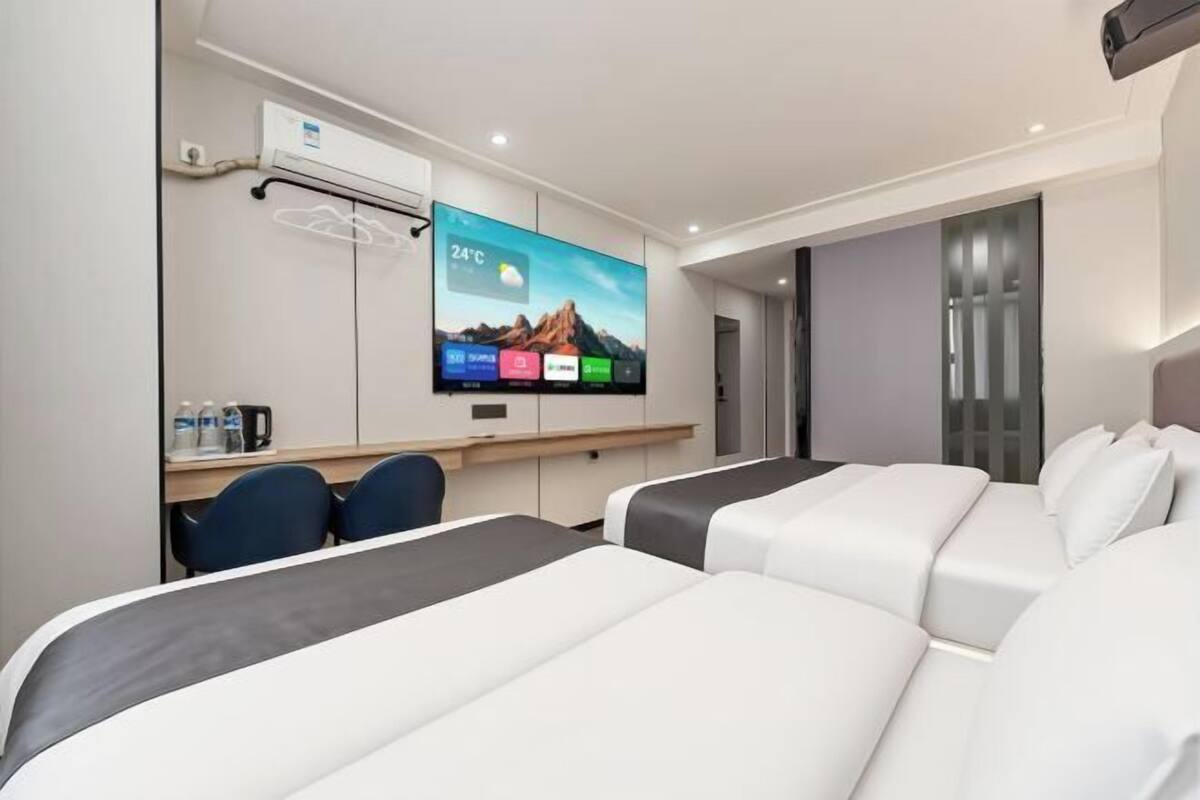 A hotel room with a bed, a desk with a computer, a TV displaying a weather forecast, and a door leading to another room.