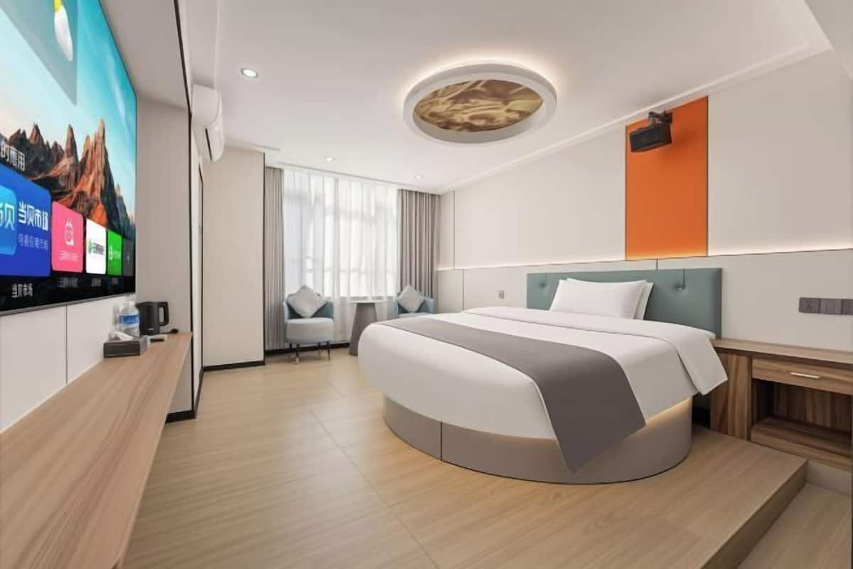 A modern hotel room with a curved bed, a flat-screen TV, a desk with a coffee maker, and a window with curtains.