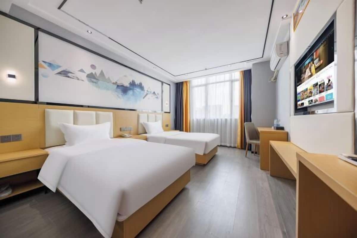 A hotel room with two beds, a desk, a chair, a TV, and a large window with curtains.