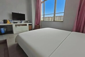 A hotel room with a bed, a desk with a flat-screen TV, a window with a view of buildings, and a trash bin.