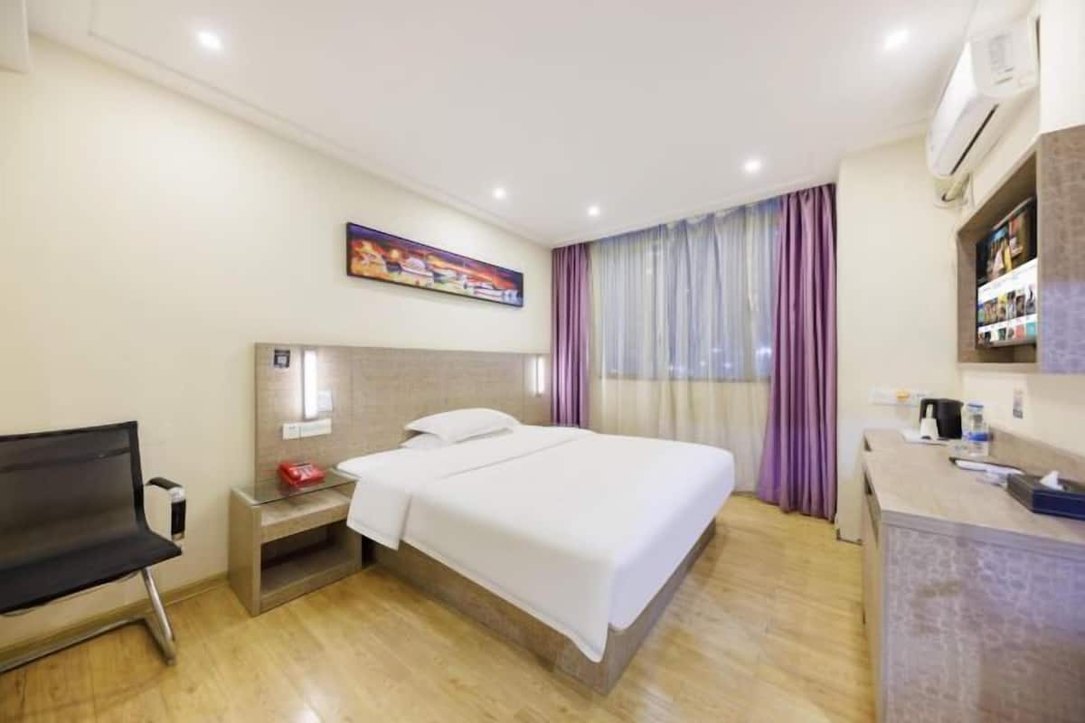 A hotel room with a bed, a desk, a chair, a nightstand, a painting, and a window with curtains.