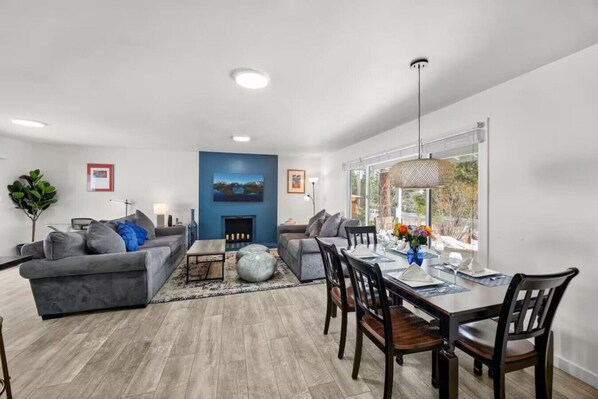 A modern living room with a fireplace, grey sofas, a dining table with chairs, and large windows offering a view.