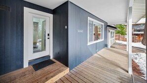 Une terrasse en bois couverte, dotée d’une porte et de fenêtres, qui donne sur une maison à l’extérieur bleu foncé.