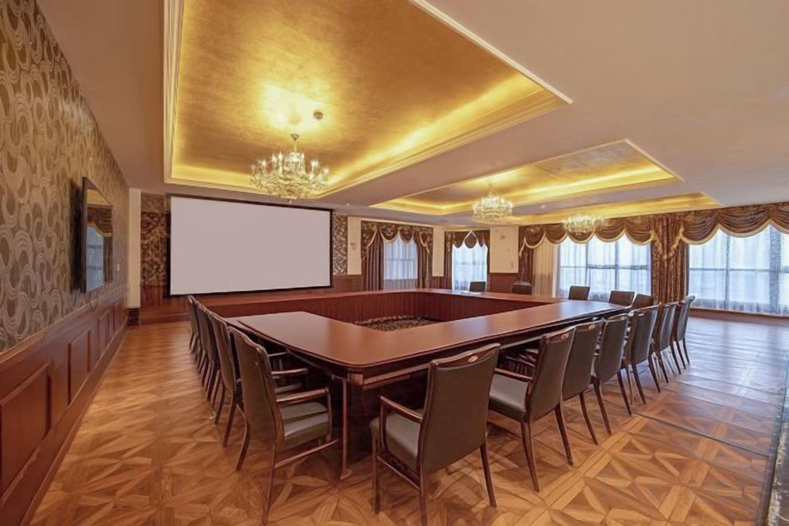 Une salle de conférence équipée d’une grande table, de chaises, d’un écran de projection et d’un lustre.