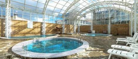 Una piscina interior con techo de cristal, rodeada de sillones de descanso.