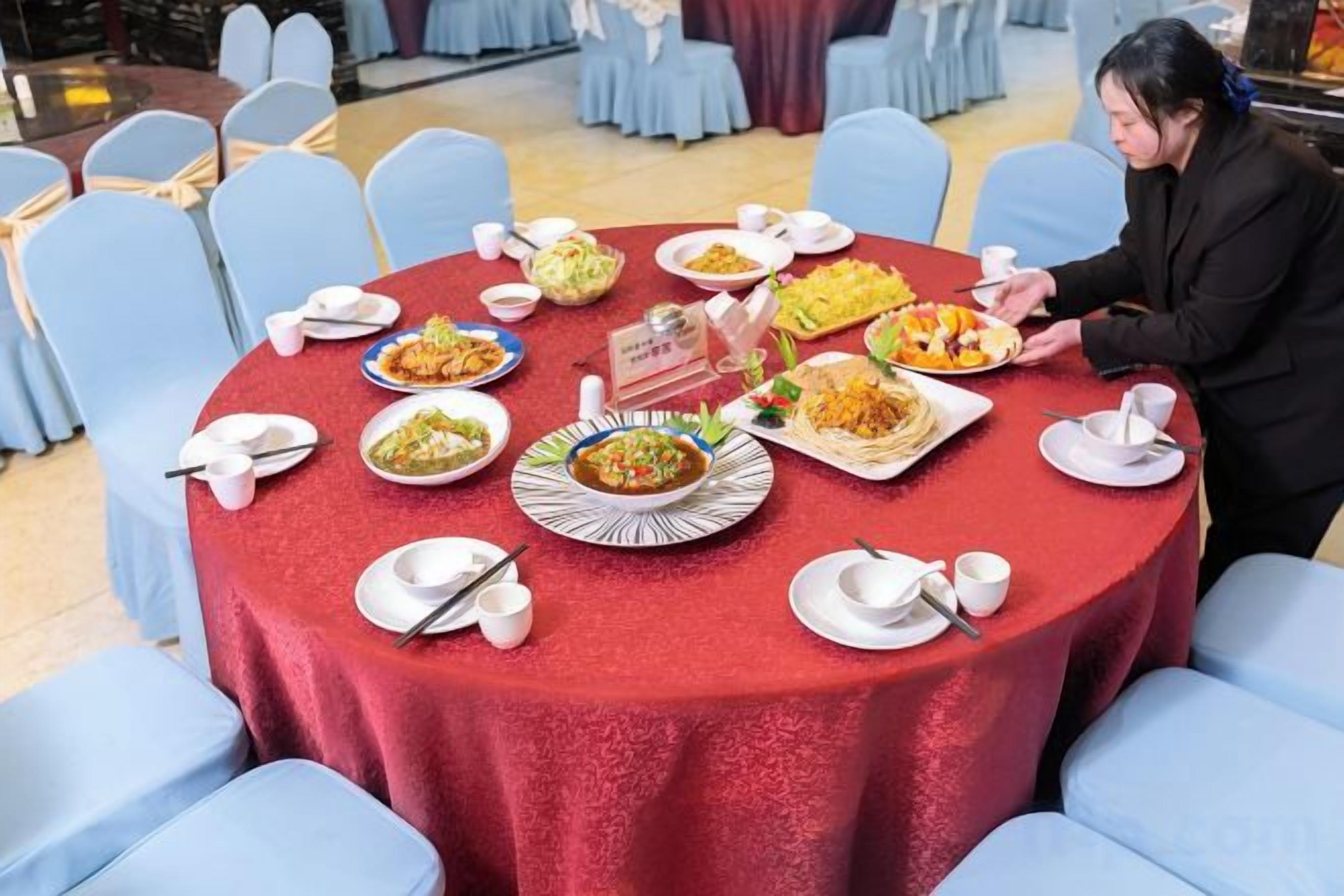 Een ronde tafel met een rode tafelkleed, omringd door blauwe stoelen, met verschillende gerechten en een persoon die eten serveert.