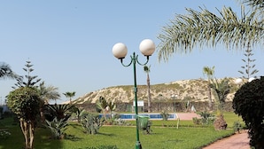 A pathway in a landscaped area with green grass, palm trees, and a mountain in the background. - Waterfront apartment (Mohammédia)