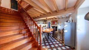 Eine Holztreppe führt zu einem Essbereich mit Schachbrettmuster, im Hintergrund ist eine Küche und rechts ein Kühlschrank zu sehen.