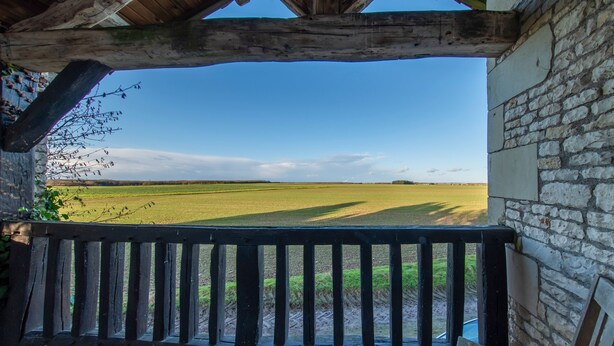 Ein hölzerner Balkon mit einer Steinsäule, der auf ein grünes Feld unter blauem Himmel blickt.