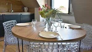 Una mesa de comedor dispuesta con platos, cubiertos y vasos, rodeada de sillas transparentes, en una habitación con techo de madera y ventana.
