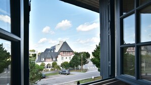 A view through a window of a house with a sloped roof and a chimney, a street with cars, and trees.