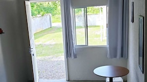 A room with a bed, a small round table, and a stool. There is a window with curtains, and a view of a grassy area outside.