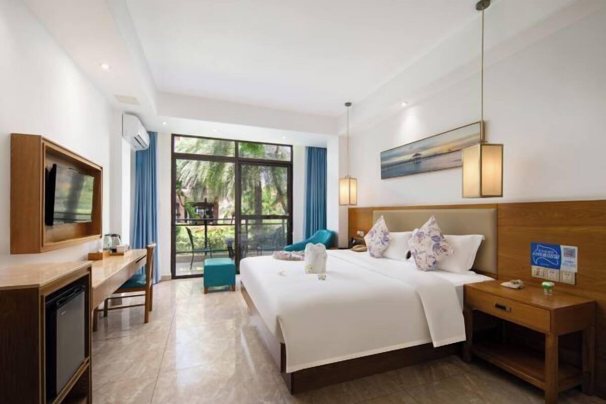 Quarto de hotel com uma cama grande, uma mesa de trabalho com televisão, uma mesinha e vista para o exterior.