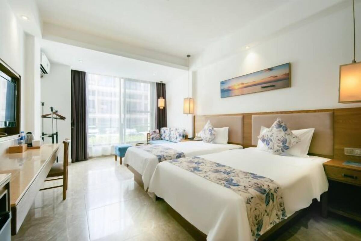 Quarto de hotel com duas camas, uma escrivaninha, uma cadeira, uma janela com cortinas, um quadro na parede e um abajur.