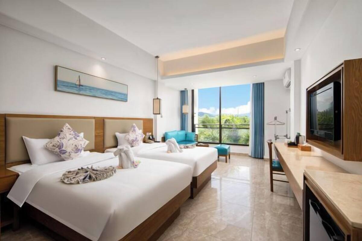 Quarto de hotel com duas camas, uma escrivaninha, uma televisão e uma janela grande com vista para o verde.