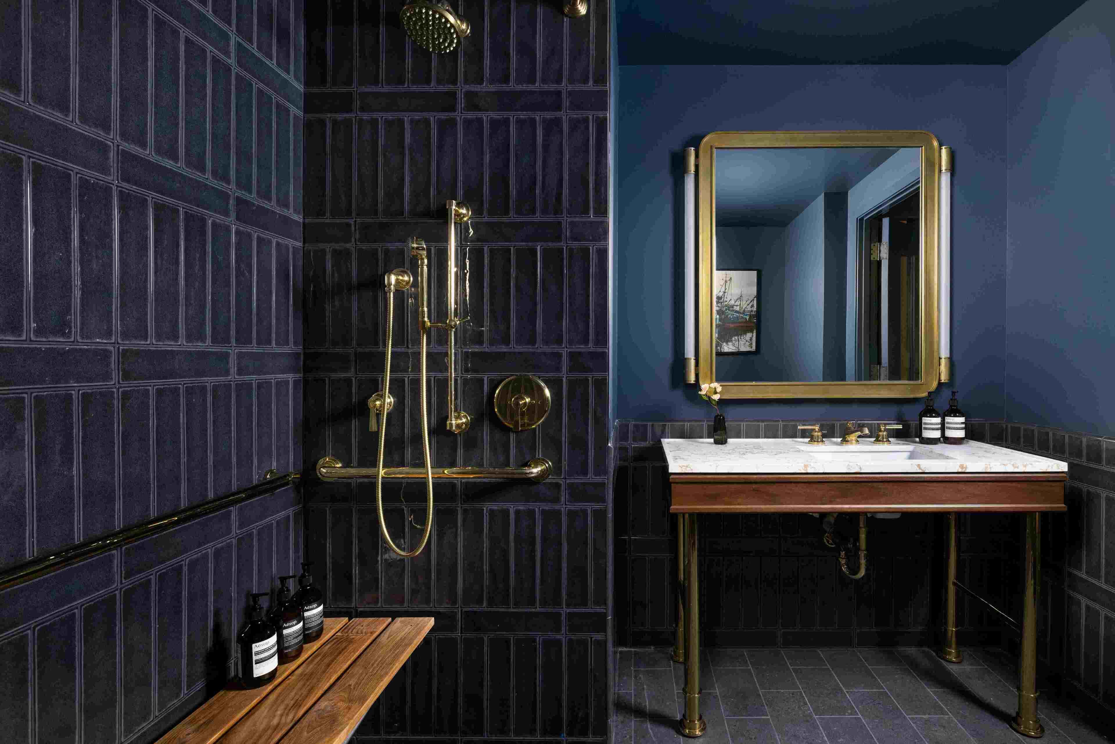 Une salle de bain aux murs recouverts de carreaux bleu foncé, avec un banc en bois, un lavabo avec un miroir et une douche dotée de robinetterie en laiton.