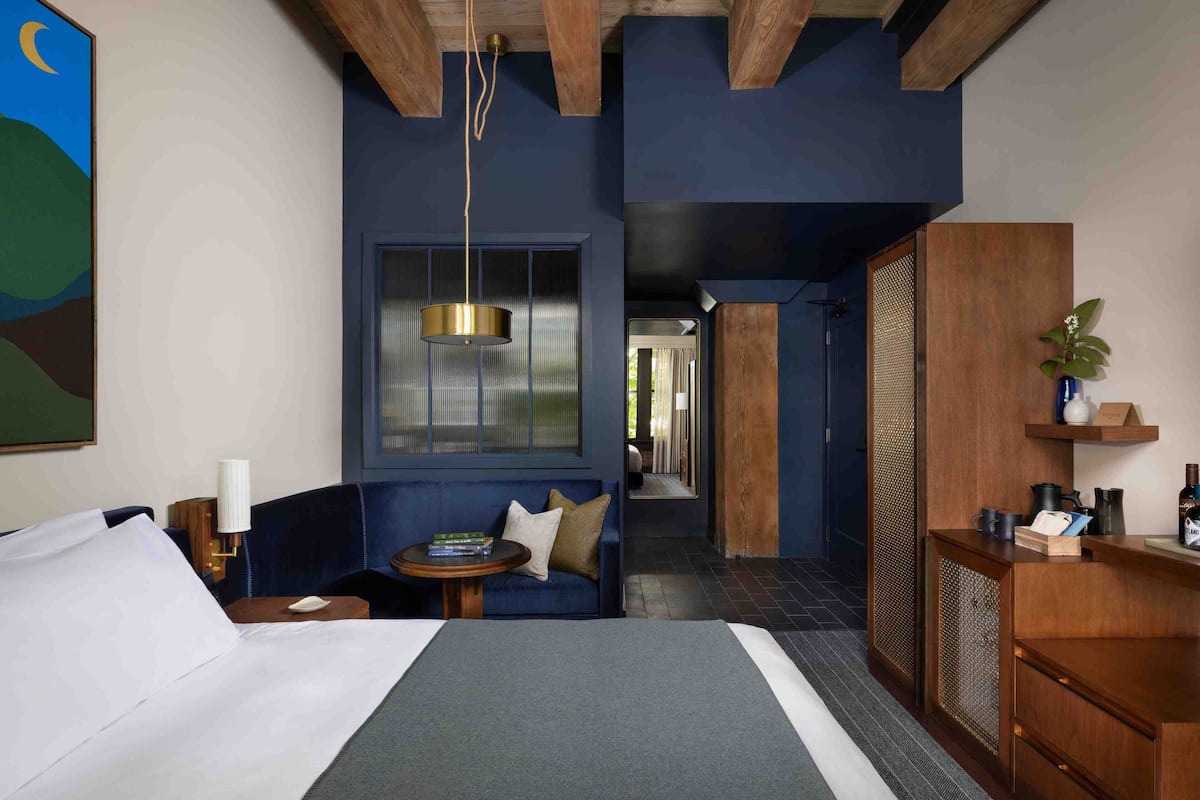 A hotel room with a large bed, a blue sofa, a small table with books, a hanging light fixture, and a wooden cabinet with a plant.