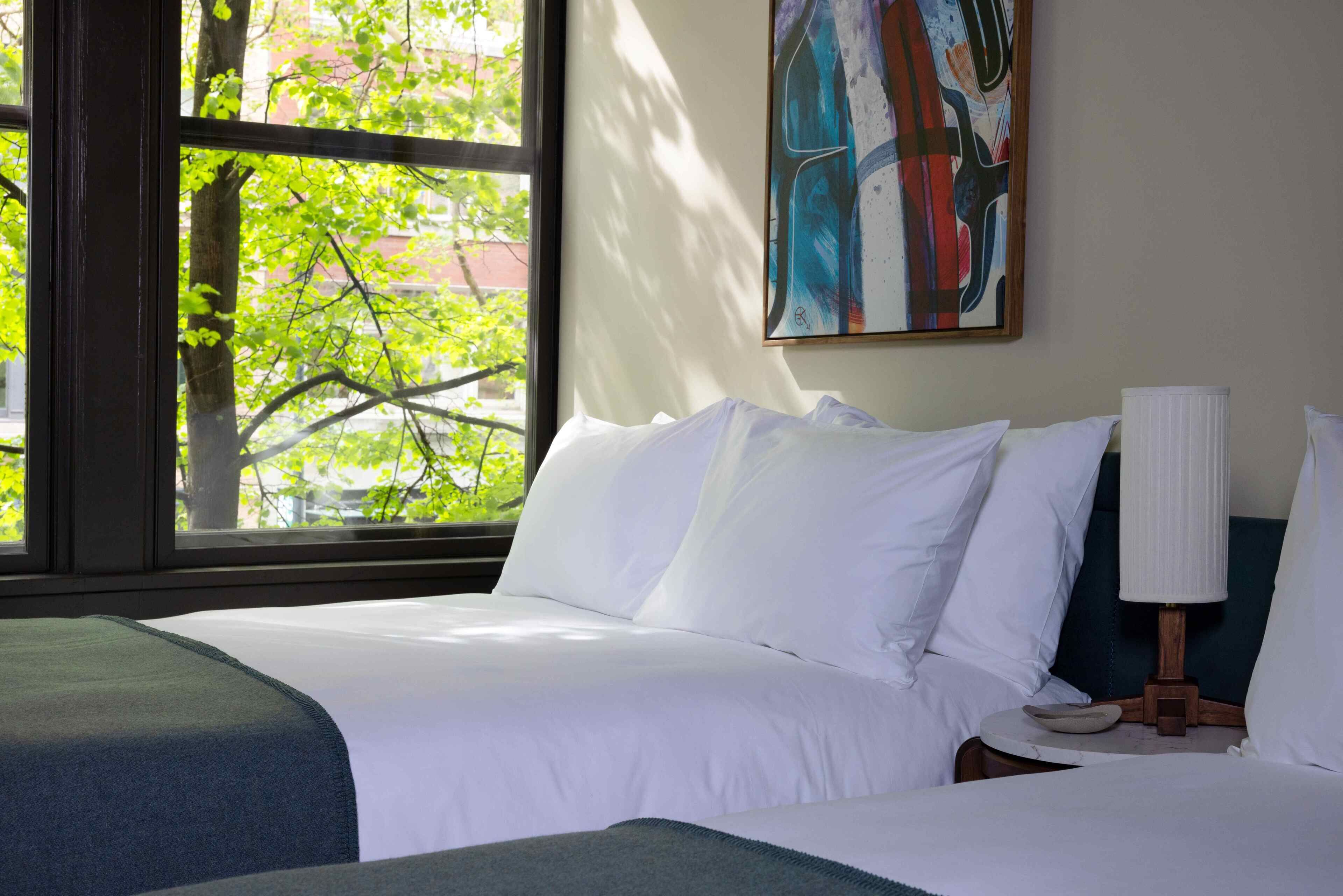 Una habitación de hotel con un ventanal grande, una cama con ropa blanca, una mesita de noche con lámpara y un cuadro en la pared.