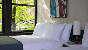 Una habitación de hotel con un ventanal grande, una cama con ropa blanca, una mesita de noche con lámpara y un cuadro en la pared.