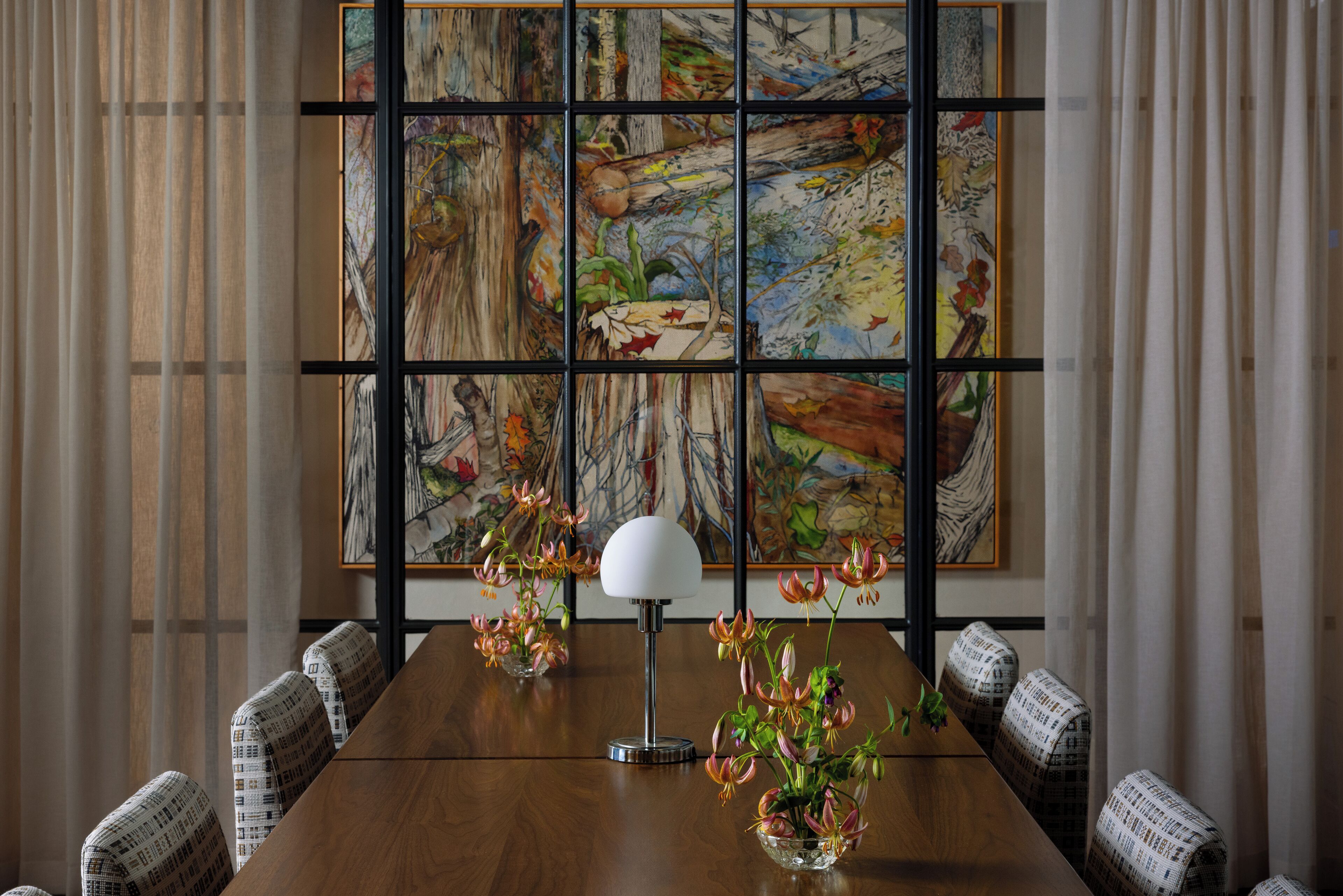 Une salle de conférence avec une table en bois, des chaises à motifs, une lampe blanche et un grand tableau abstrait accroché au mur.