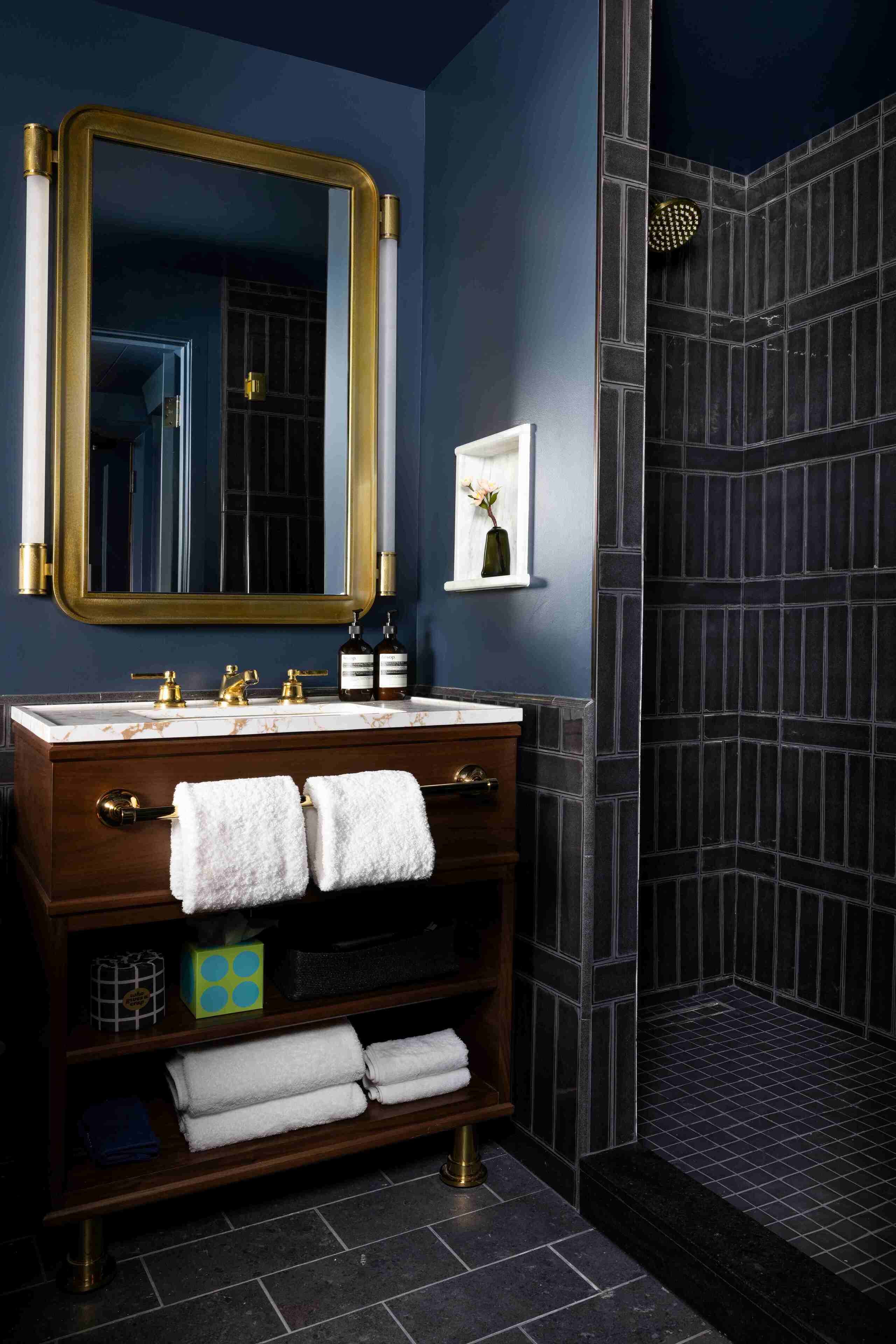 Une salle de bain avec un mur bleu foncé, un miroir encadré d’or, un meuble-lavabo en bois surmonté d’un plan de travail en marbre et une douche aux carreaux foncés.