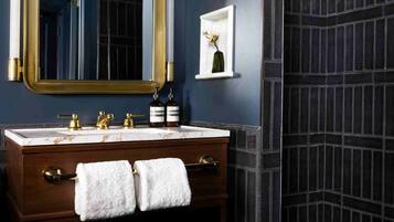 Une salle de bain avec un mur bleu foncé, un miroir encadré d’or, un meuble-lavabo en bois surmonté d’un plan de travail en marbre et une douche aux carreaux foncés.