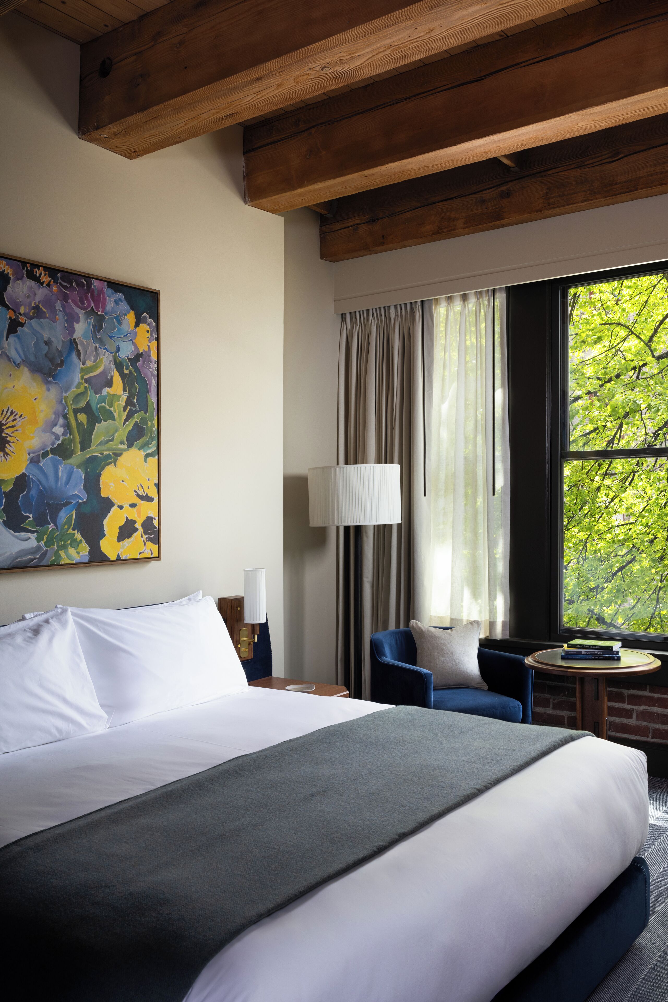 A hotel room with a large bed, a blue chair, a small round table, and a window with a view of trees.