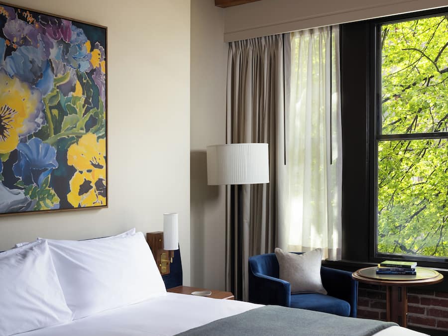Habitación de hotel con una cama grande, una silla azul, una mesita redonda y una ventana con vistas a árboles.