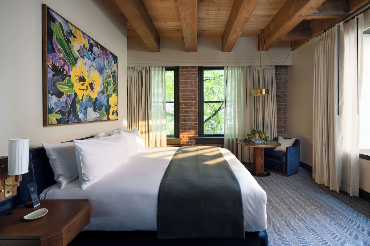 A hotel room with a large bed, a painting on the wall, a window with curtains, a small table with a chair, and a lamp on a bedside table.