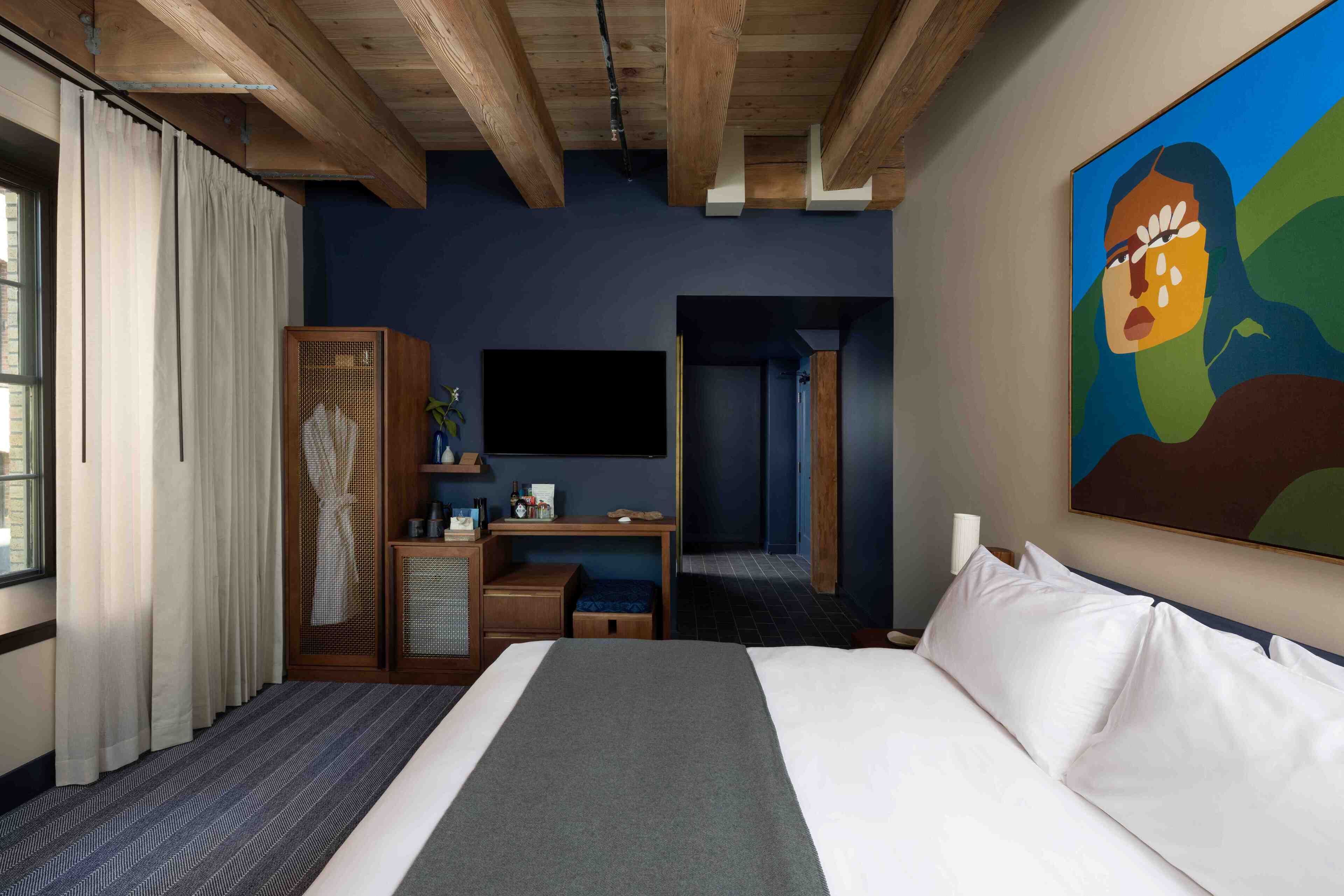A hotel room with a bed, a desk, a TV, and a painting on the wall.