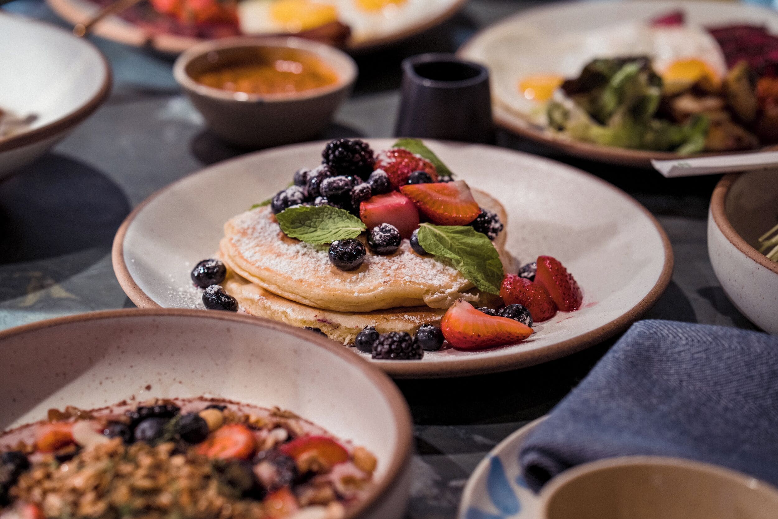A variety of breakfast dishes on plates, including pancakes topped with berries and powdered sugar.