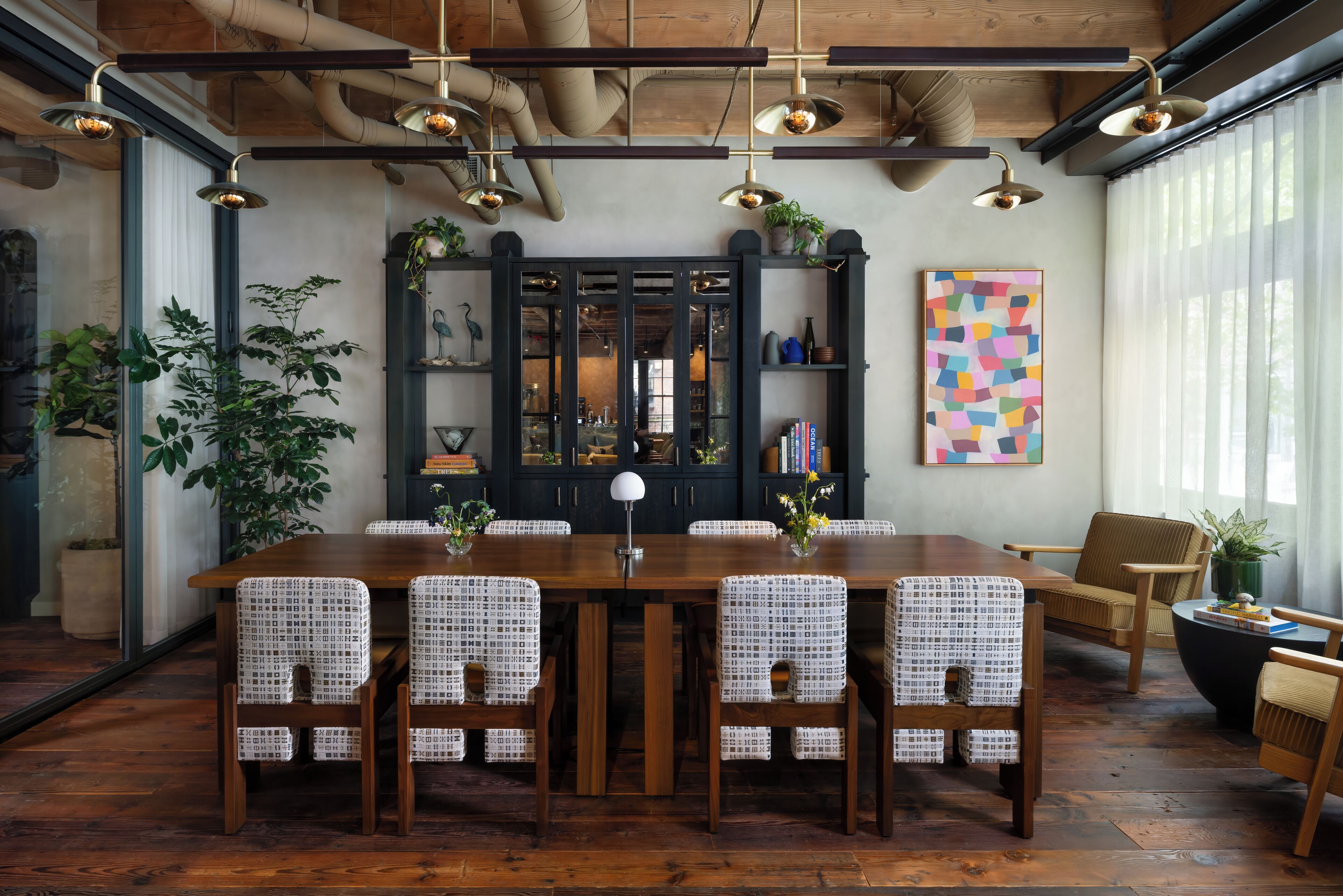 A dining area with a wooden table and patterned chairs, a dark wooden cabinet with glass doors, and a colorful abstract painting on the wall.