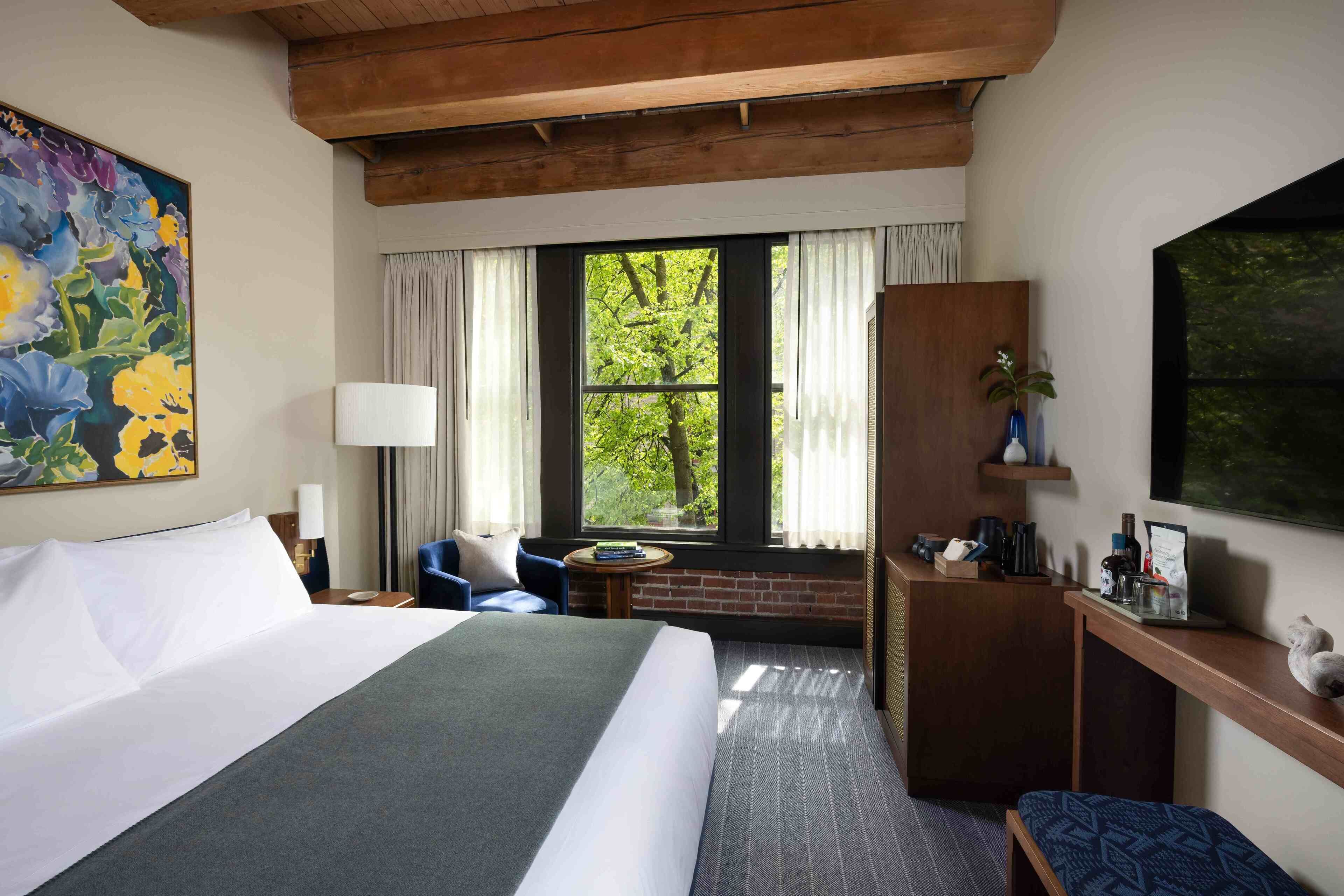 A hotel room with a large bed, a desk, a chair, a TV, and a window with a view of trees.