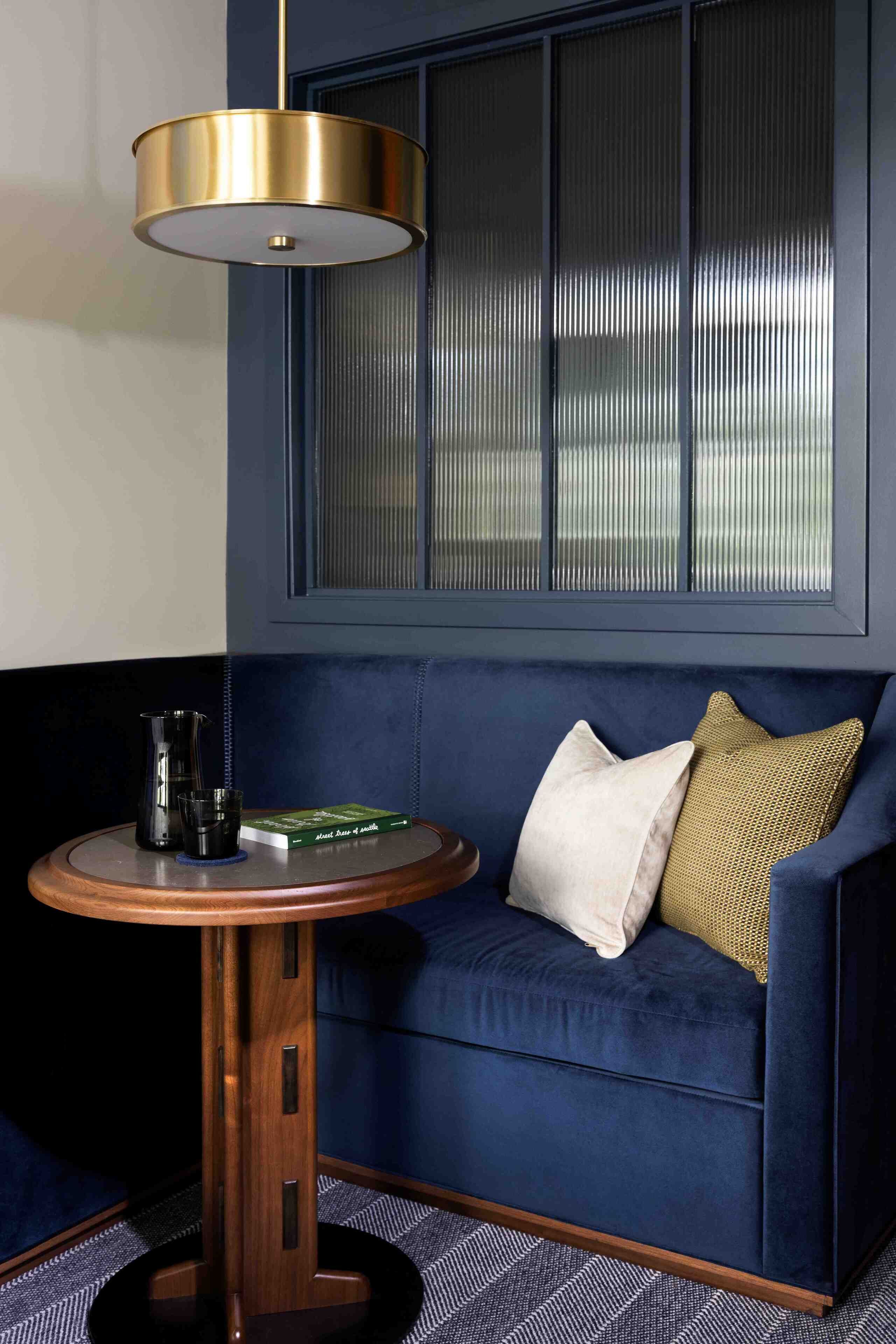 Una habitación de esquina con un sofá azul oscuro, una mesa redonda de madera y un objeto negro, además de una lámpara colgante con marco dorado.