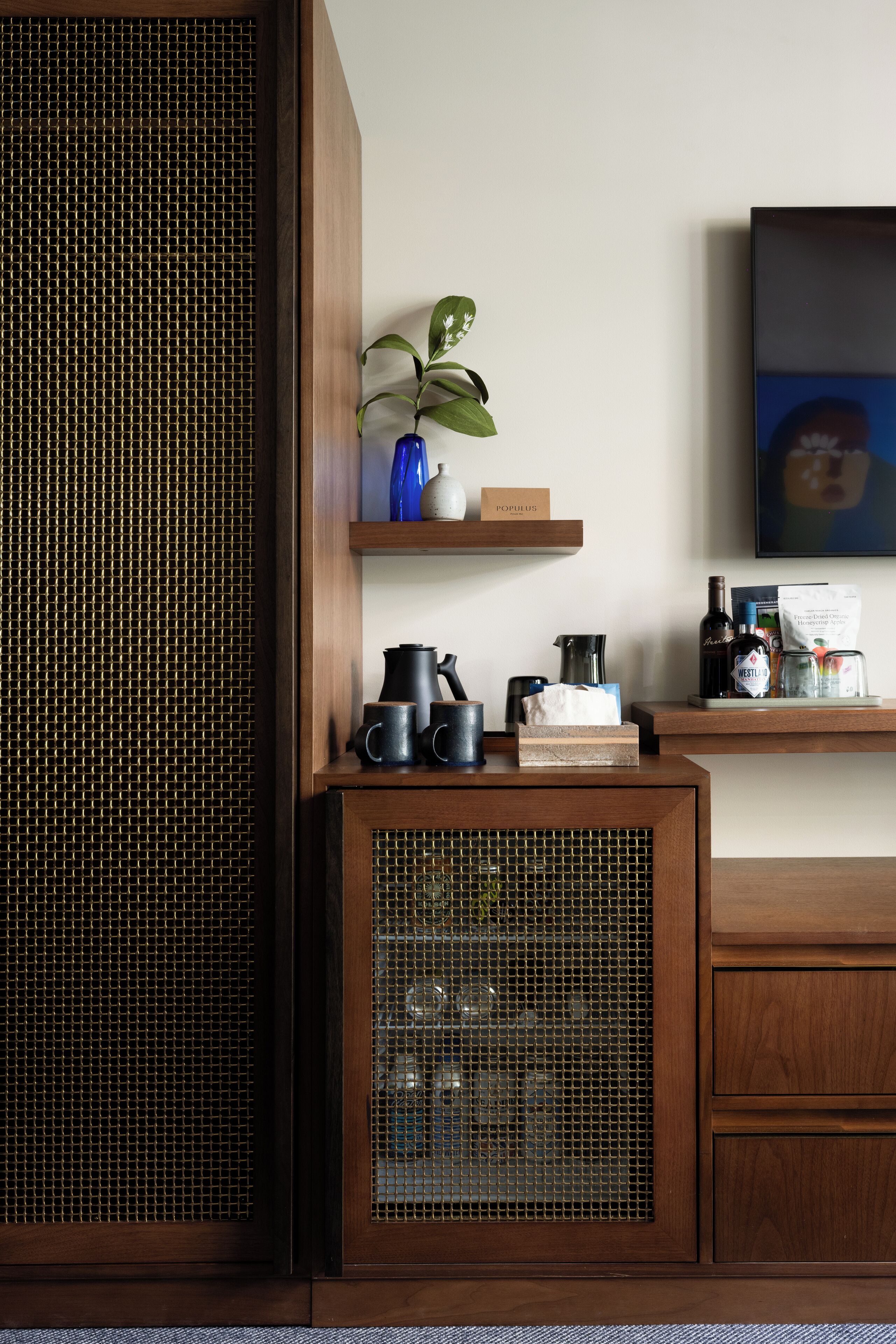 Un gabinete de madera con frente de malla, una estantería con una planta, una cafetera y un cuadro en la pared.