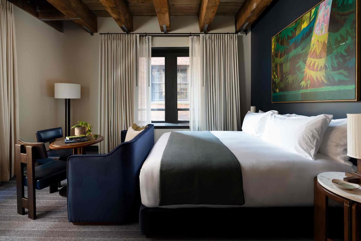 A hotel room with a large bed, a desk with a lamp, a chair, a small table with a plant, and a painting on the wall.