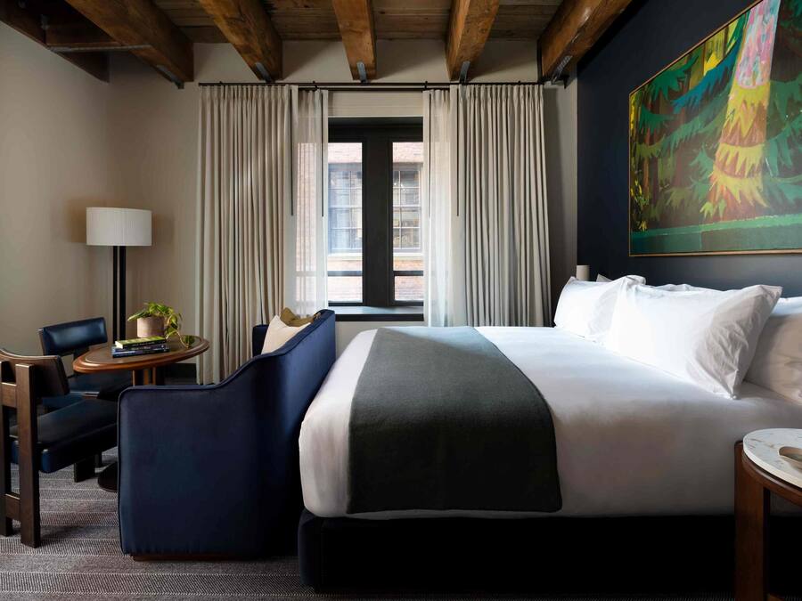 A hotel room with a large bed, a desk with a lamp, a chair, a small table with a plant, and a painting on the wall.
