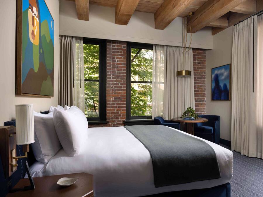 Una habitación de hotel con una cama grande, un escritorio con lámpara, una silla, una mesita con una planta y un cuadro en la pared.