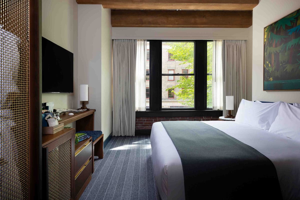 A hotel room with a large bed, a desk with a flat-screen TV, a window with curtains, and a painting on the wall.